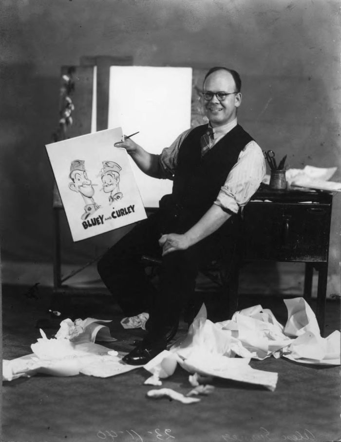Early war-time studio portrait of Alex Gurney introducing ‘Bluey and Curley’ as soldiers, the waste paper around his feet deliberately left to indicate the time and care he took with his drawings. (Taken November 1940, photographer unknown. Reproduced courtesy of Vane Lindesay) [black and white photograph]